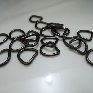 Könnte beinhalten: Eine Gruppe schwarzer D-Ringe aus Metall, auch bekannt als D-Ringe oder D-förmige Ringe. Diese Ringe werden häufig im Handwerk, beim Nähen und bei der Schmuckherstellung verwendet.