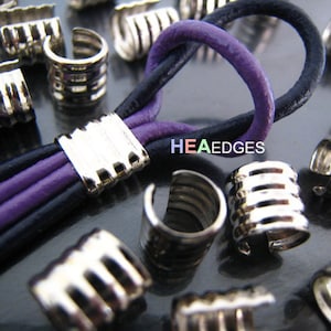 Puede incluir: Terminales de cordón de metal plateado con acabado texturizado. Los terminales de cordón se muestran con cordón de cuero negro y morado. La imagen también muestra una variedad de terminales de cordón en una pila.