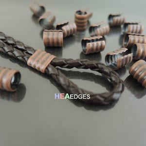 Puede incluir: Primer plano de un conjunto de terminales de cordón de metal de color cobre. Las terminales tienen un diseño acanalado y están esparcidas sobre una superficie oscura. Un cordón de cuero trenzado está enhebrado a través de dos de las terminales.