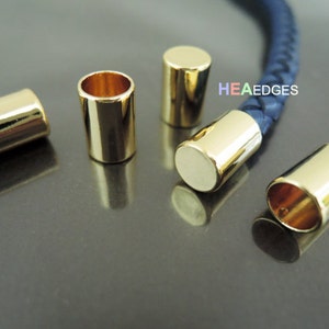 Puede incluir: Cinco tapones de cordón de metal de color dorado. Los tapones son cilíndricos y tienen un acabado liso y brillante. Los tapones se muestran en un cordón de cuero azul.