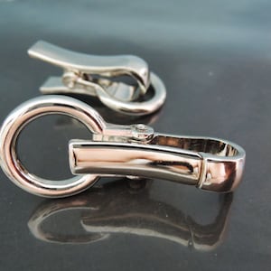 May include: A silver metal clasp with a round ring and a hook. The clasp is designed for attaching to a strap or chain.