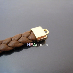 May include: Close-up of a brown leather braided bracelet with a gold metal clasp. The clasp is a square shape with a small loop on the top.