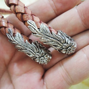 May include: A close-up of a leather cord bracelet with two silver floral-shaped end caps. The bracelet is braided and has a light brown color.