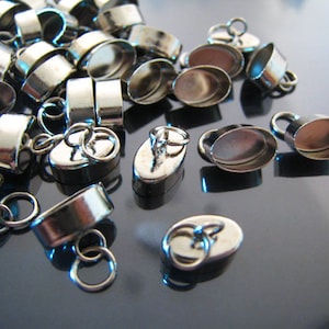 May include: A group of silver oval-shaped pendant caps with a loop on top. The caps are designed to hold a small piece of glass or other material.