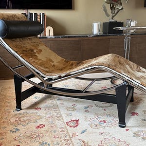 May include: A modern chaise lounge with a cowhide seat and a black leather headrest. The frame is made of chrome and black metal. The chair is placed on a patterned rug.