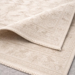 Könnte beinhalten: Ein cremefarbener Teppich mit einem dezenten, tonalen Blumenmuster und komplexen botanischen Motiven. Der Teppich hat eine dekorative Bordüre und eine kurze, geknotete Franse am Rand, wobei eine Ecke umgeschlagen ist, um die Textur zu zeigen.