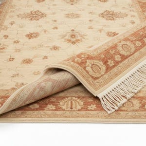 May include: Beige and rust-colored area rug with a floral pattern. The rug is partially rolled up, revealing the underside and fringe. The design features repeating floral motifs and a border with a detailed pattern.