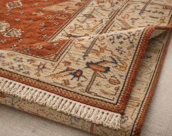Puede incluir: Una alfombra rectangular con un campo central de color rojo óxido profundo con intrincados motivos geométricos y florales en crema, azul y marrón. La alfombra tiene un borde ancho de color crema con patrones detallados y flecos cortos.