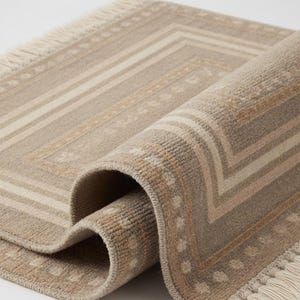 Puede incluir: Alfombra rectangular beige y marrón claro con un patrón geométrico y flecos. La alfombra está parcialmente enrollada, revelando la parte inferior y el diseño detallado. El patrón incluye rayas y pequeños motivos florales.