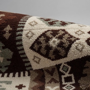 Könnte beinhalten: Nahaufnahme eines aufgerollten, gemusterten Teppichs. Der Teppich weist ein sich wiederholendes geometrisches Design in Braun-, Creme- und gedeckten Grüntönen auf. Die Textur wirkt weich und flauschig, mit detaillierter Weboptik. Das Design umfasst Diamanten und Rechtecke.