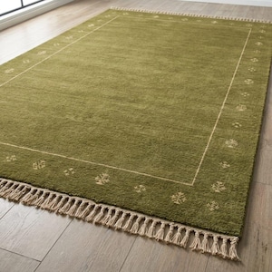 May include: A rectangular olive green area rug featuring a thin cream-colored border line and a repeating small floral pattern along the perimeter. The rug has a textured pile surface and is finished with long, cream-colored fringe tassels on both ends.