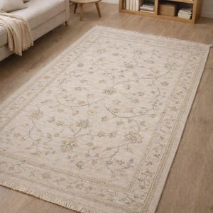 Op de afbeelding: Beige rechthoekig vloerkleed met een bloemenpatroon en gefranjerde randen. Het vloerkleed heeft een gedetailleerd ontwerp van bloemen en wijnranken in beige, groen en bruin, geschikt voor een woonkamer.