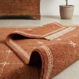 May include: A rectangular area rug in a deep terracotta orange color featuring a white grid pattern with small star-like accents at the intersections. The rug has a textured pile surface, a decorative border, and short fringe along the edge.