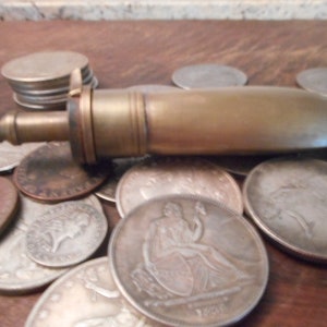May include: A brass, cylindrical, antique-style,  decorative bottle stopper with a small, round, brass cap on one end. The stopper is lying on a surface covered with silver coins.