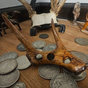 May include: A close-up of a deer antler with a series of holes drilled through it. The antler is resting on a wooden surface with a pile of silver coins scattered around it. The antler is a light brown color with a darker brown stain on the top.