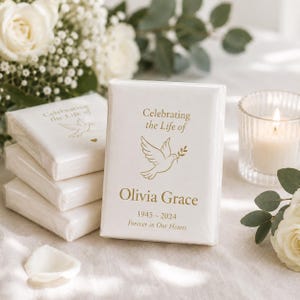 May include: White memorial favors with gold text and a dove illustration. The text reads "Celebrating the Life of Olivia Grace 1945-2024 Forever in Our Hearts." A lit candle and white flowers are in the background.
