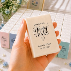 以下が含まれることがあります： 「FOR YOUR Happy TEARS」の黒い筆記体とハートのシンボルが描かれた、小さなオフホワイトの長方形のパッケージ。パッケージには「Luna & Ethan」と日付「29.08.2026」も含まれています。背景には他のパッケージが見えます。