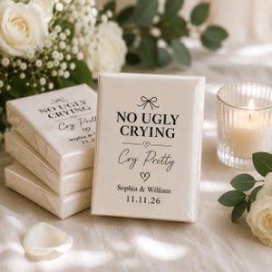 May include: Cream-coloured tissue boxes with the text "NO UGLY CRYING" and "Cry Pretty" in black script. The boxes are stacked near a lit candle and white roses, with the names Sophia & William and the date 11.11.26 printed on the front.
