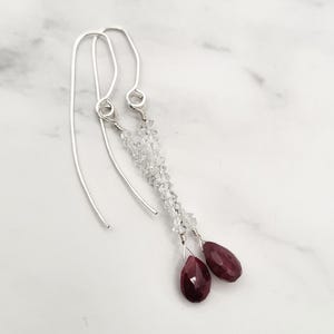 May include: Dangle earrings with silver hooks, clear crystal beads, and two faceted, teardrop-shaped, deep red gemstones. The earrings are displayed on a white marble surface.