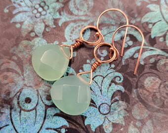 May include: A pair of earrings featuring pale green, faceted teardrop-shaped gemstones. The gemstones are suspended from copper-colored wire and ear hooks. The earrings are displayed against a patterned background with teal and dark gray floral designs.