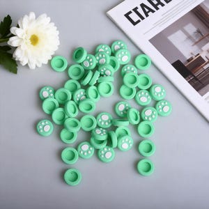 May include: A collection of mint green joystick thumb grips with white paw prints. The grips are scattered on a light gray surface, with a white flower and a magazine in the background. These grips are designed for gaming controllers.