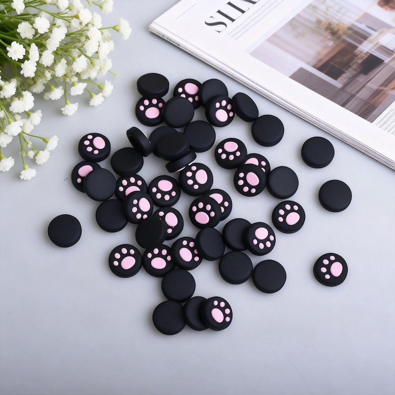 Puede incluir: Un conjunto de tapas de joystick negras y circulares con huellas de patas rosas. Algunas tapas son negras lisas. Los art&iacute;culos est&aacute;n dispersos sobre una superficie gris claro, con una revista y flores blancas en el fondo.