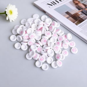 May include: A collection of white, circular controller thumb grips with pink paw prints. The grips are scattered on a light gray surface, with a white flower and a magazine in the background. These grips are designed for gaming.