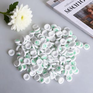 May include: A pile of white, circular controller thumb grips with a light green paw print design. The grips are made of a smooth material and are scattered on a light gray surface. A white flower and a magazine are in the background.
