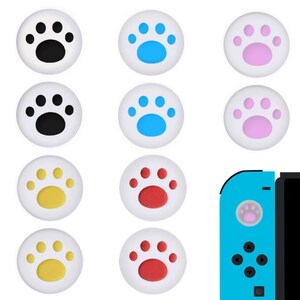 Puede incluir: Un conjunto de tapas de joystick blancas con dise&ntilde;os de huellas de patas de colores. Las huellas de patas son negras, azules, rosas, amarillas y rojas. Se muestra un mando de juego azul con una tapa rosa.