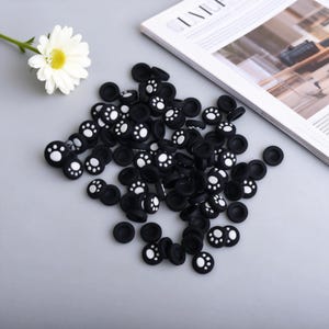 May include: A collection of black joystick thumb grips with white paw print designs. The grips are scattered on a light gray surface, with a white daisy and a magazine in the background.