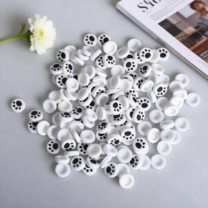 May include: A collection of white, circular controller thumb grips with black paw print designs. The grips are scattered on a light gray surface, with a white flower and a magazine visible in the background. These grips are designed for gaming.