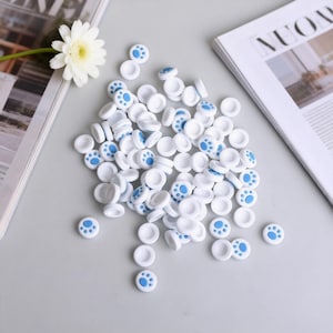 May include: A collection of white, circular buttons with blue paw print designs. The buttons are scattered on a light gray surface, with a white flower and a magazine in the background. The buttons are likely for a craft or DIY project.