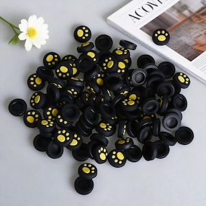 May include: A collection of black joystick thumb grips with yellow paw prints. The grips are scattered on a light gray surface, with a white daisy and a magazine in the background. The grips are designed for video game controllers.