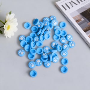 May include: A collection of light blue joystick thumb grips with white paw print designs. The grips are scattered on a light gray surface, with a magazine and a white flower in the background. These are designed to enhance gaming controller aesthetics and grip.