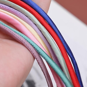 May include: A set of eight satin ribbon cords in various colours, including red, blue, green, purple, pink, and cream. The cords are arranged in a rainbow pattern and are held in a hand.
