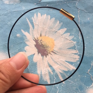 Könnte beinhalten: Ein schwarzes elastisches Armband mit einem goldenen Verschluss. Das Armband wird in einer Hand gehalten und befindet sich vor einem Vintage-Blumendruck.