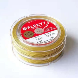 Puede incluir: Un carrete de alambre dorado recubierto de nailon, etiquetado como "FLEXY7", con una etiqueta roja y blanca. La etiqueta indica que el alambre tiene 0,40 mm de diámetro y 100 m de longitud. El alambre está diseñado para la fabricación de joyas y manualidades.