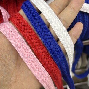 Può includere: Primo piano di un bordo intrecciato colorato in rosa, rosso, blu e bianco. Il bordo è realizzato in un materiale morbido e tessuto ed è disposto in una mano. Il design intrecciato è visibile, mostrando la consistenza e i dettagli del bordo.