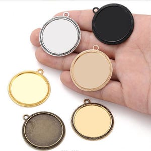 May include: A collection of round pendant trays in various metallic finishes: silver, gold, bronze, and black. Each tray has a circular inset and a small loop for attaching a chain or cord. The trays are designed for jewelry making.