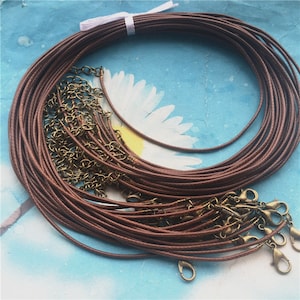 Może przedstawiać: Pakiet hurtowy brązowego woskowanego sznurka, około 20 nici, ze złotymi karabińczykami. Sznurek idealnie nadaje się do tworzenia naszyjników, bransoletek i innych ozdób.