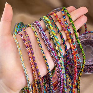 Peut inclure: Plusieurs brins de cordon tressé coloré sont présentés dans une main. Les cordons sont de différentes couleurs, notamment jaune, bleu, rouge et vert. Les cordons sont probablement utilisés pour la fabrication de bijoux ou d'autres travaux manuels.