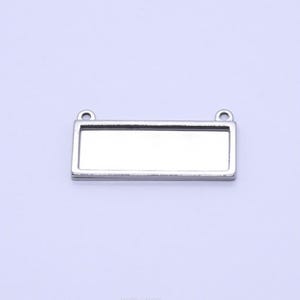 Puede incluir: Un colgante rectangular plateado con una superficie clara y reflectante. El colgante tiene dos pequeños bucles en la parte superior para sujetarlo a una cadena u otros componentes de joyería. El engaste está diseñado para sostener un inserto rectangular.