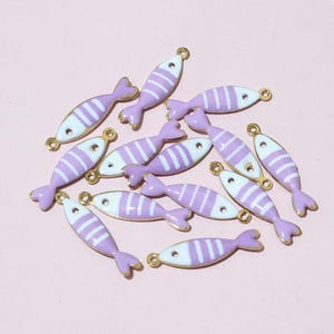 May include: A collection of small, fish-shaped charms. Each charm features a lavender body with white stripes, a lavender tail, and gold-coloured accents. The charms are scattered on a light pink surface.