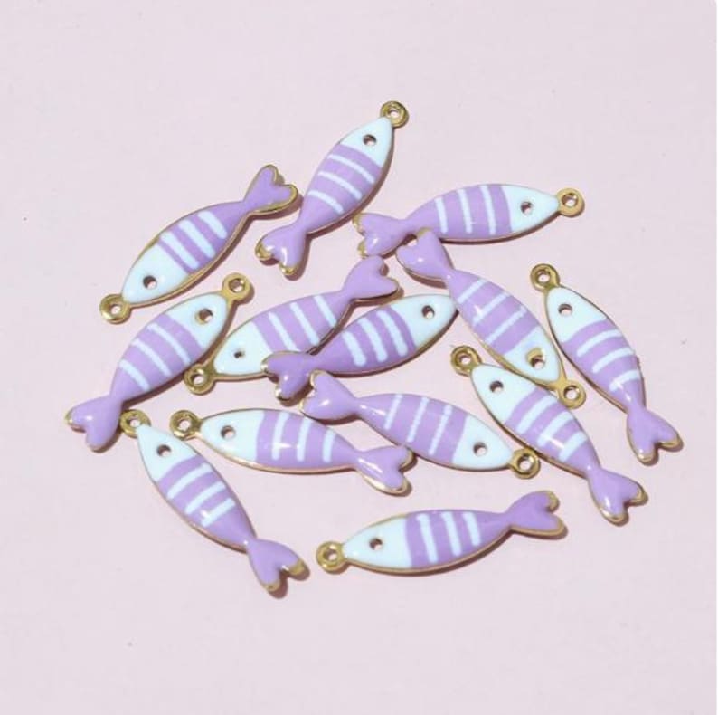 May include: A collection of small, fish-shaped charms. Each charm features a lavender body with white stripes, a lavender tail, and gold-coloured accents. The charms are scattered on a light pink surface.