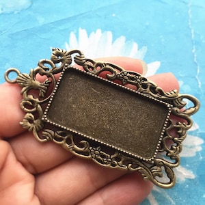 Könnte beinhalten: Ein antikes rechteckiges Anhänger-Tablett in Bronzefarbe mit einem verzierten Filigranrahmen. Das Tablett hat einen rechteckigen Einsatz für einen Cabochon oder eine andere Verzierung. Der Rahmen weist florale und scrollartige Designs sowie eine kleine Schlaufe zum Aufhängen auf.