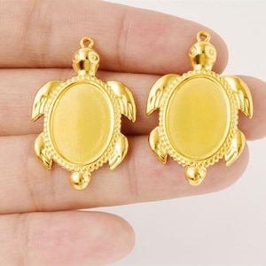 May include: Two gold-colored turtle-shaped pendants. Each pendant has an oval center, a turtle head, and flipper-like wings. The pendants are made of a shiny, reflective metal.