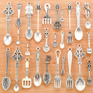 May include: A collection of 20 silver-toned metal charms in the shape of spoons, forks, and other kitchen utensils. The charms are arranged on a wooden surface.