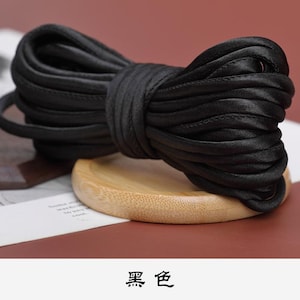 May include: A black satin cord, coiled and tied into a bow shape, resting on a wooden surface. The cord is approximately 1.2 cm wide.