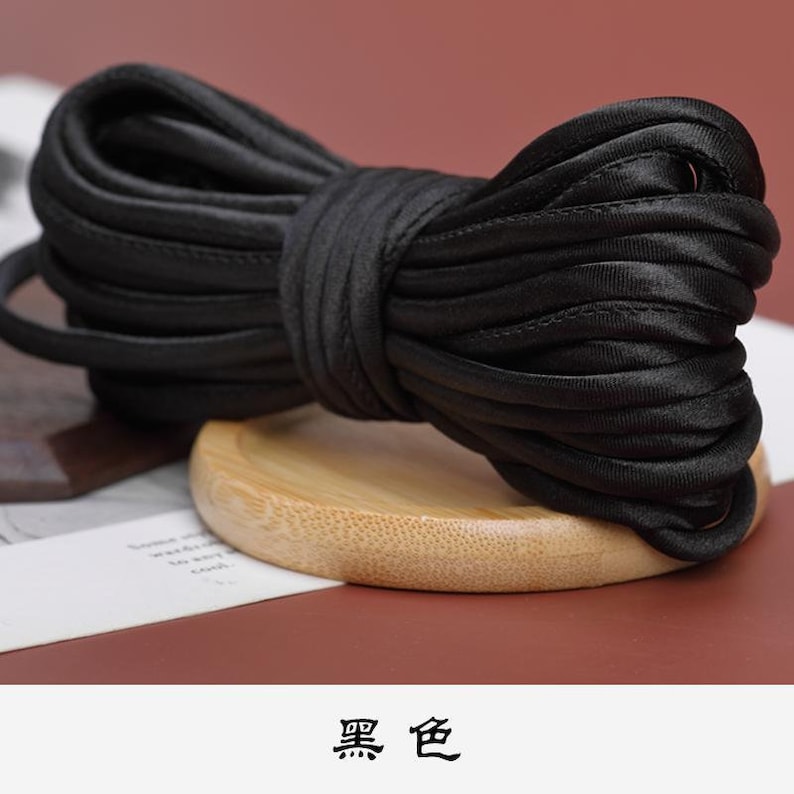 May include: A black satin cord, coiled and tied into a bow shape, resting on a wooden surface. The cord is approximately 1.2 cm wide.