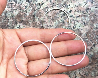 Puede incluir: Primer plano de tres círculos delgados de metal plateado. Dos círculos se sostienen en una mano, mientras que el tercero está posicionado encima. Los círculos son simples, minimalistas y parecen ser componentes de joyería.